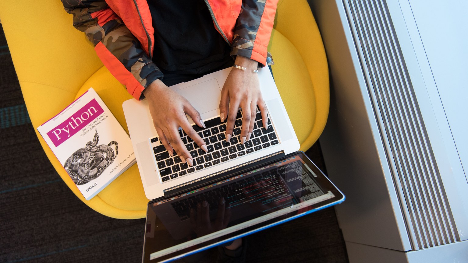 High-angle view of woman coding on a laptop, with a Python book nearby. Ideal for programming and tech content.